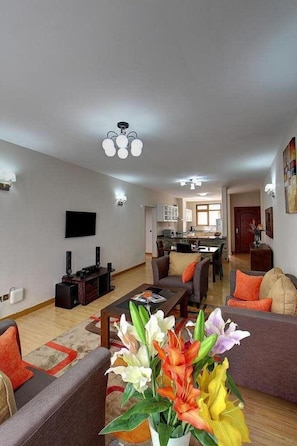 A modern living room with a flat-screen TV, a coffee table, and a sofa. There is a kitchen in the background with cabinets and a dining area. - Fenesi Gardens Apartments (Nairobi)