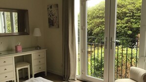 A hotel room with a bed, a desk, a chair, a balcony with a view of greenery, and a window with curtains. - Kilchurn Suites (Dalmally)