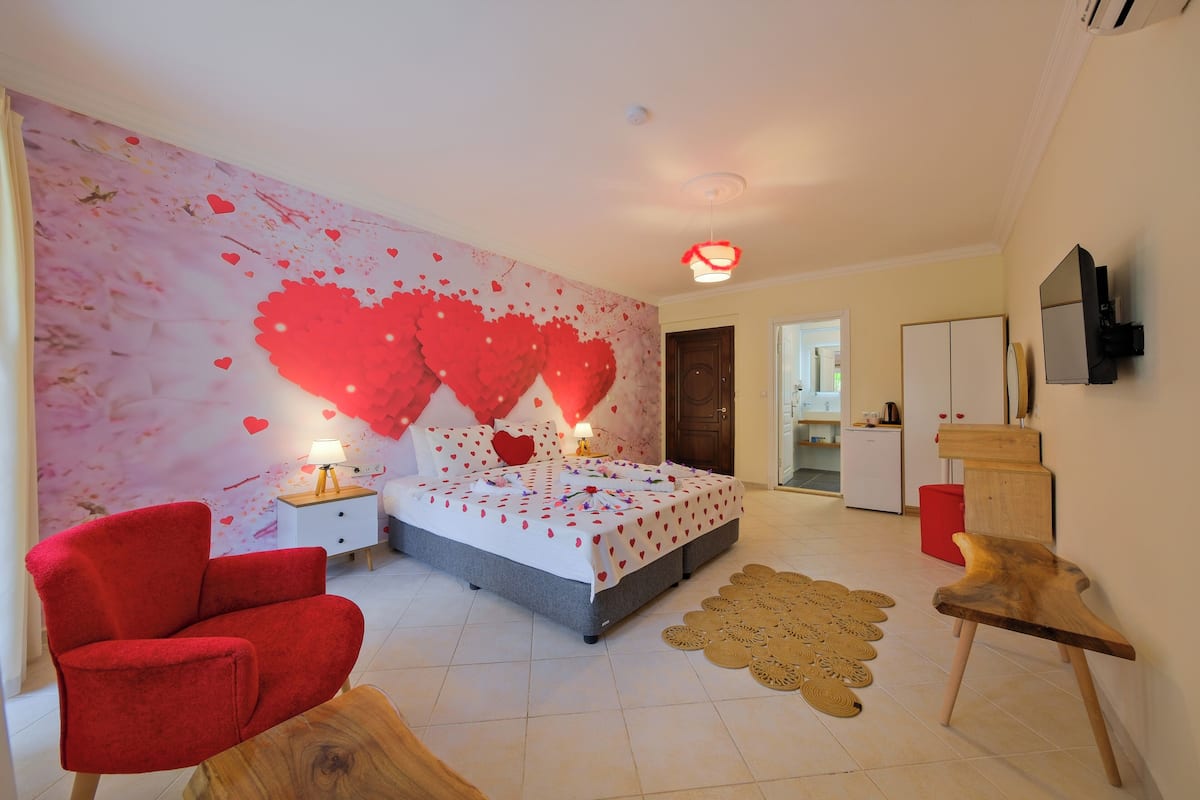 A bedroom with a bed, a red armchair, a wooden side table, a wall-mounted TV, and a kitchen area in the background.