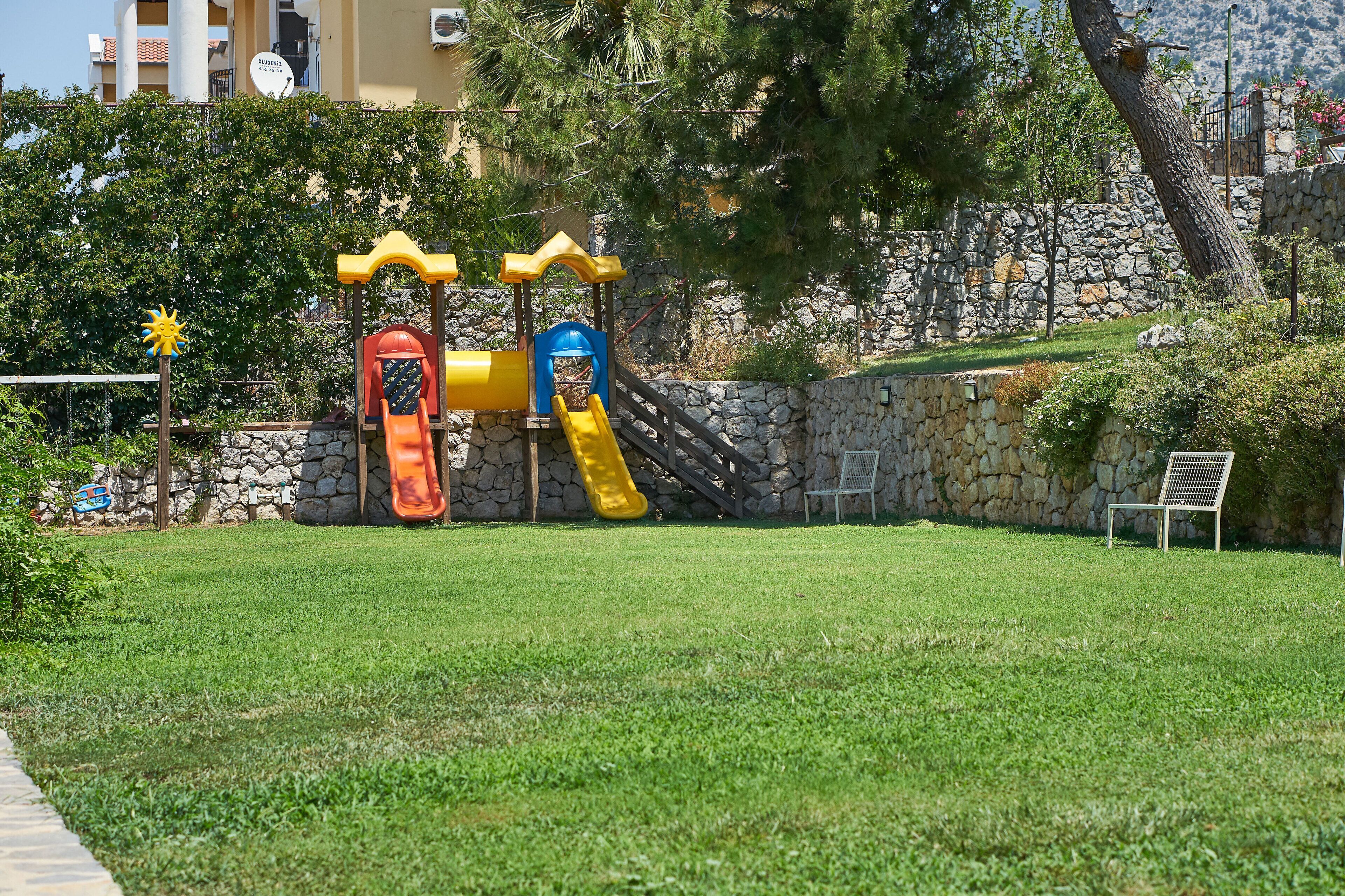 A playground with slides and climbing structures, surrounded by a stone wall and greenery.