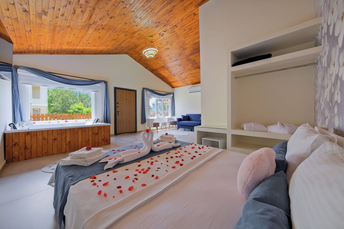 A bedroom with a wooden ceiling, a large bed with rose petals, a bathtub, and a view of greenery outside.