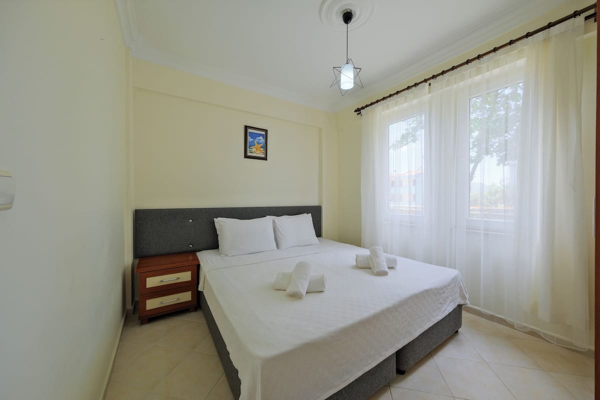 A neatly made bed with white linens and two towels, a bedside table with a lamp, and a window with sheer curtains.