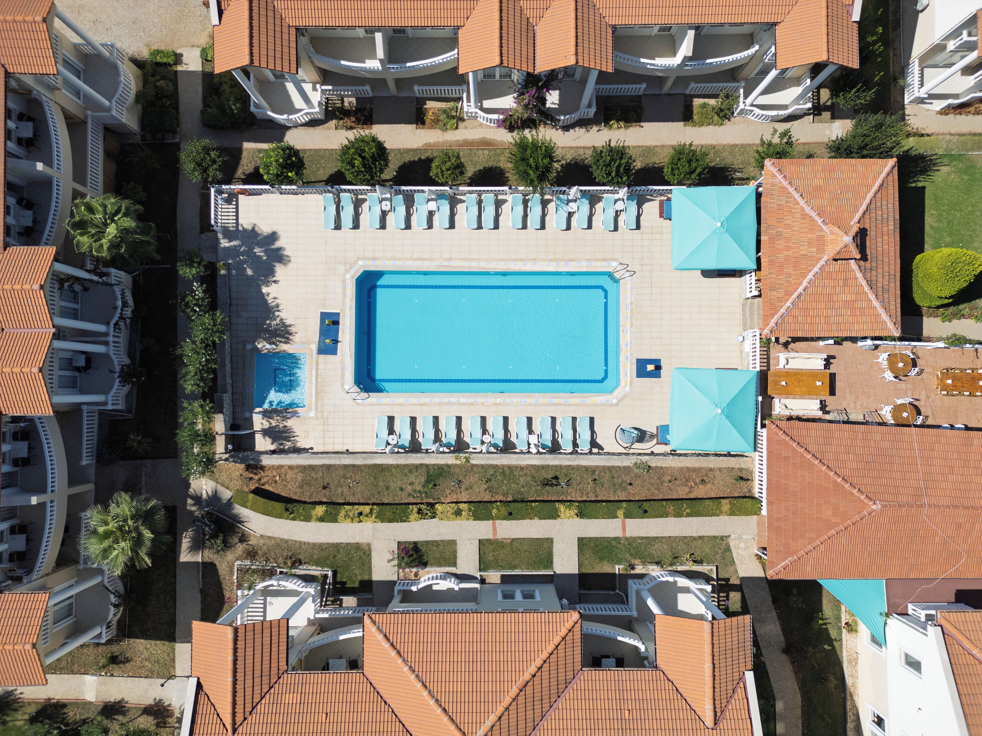 An aerial view of a hotel with a central swimming pool surrounded by lounge chairs and umbrellas, adjacent to a landscaped garden.