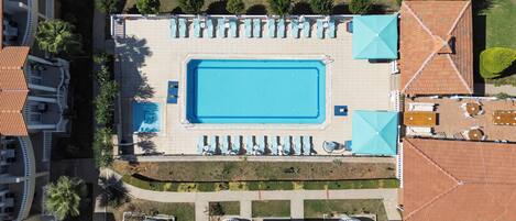 An aerial view of a hotel with a central swimming pool surrounded by lounge chairs and umbrellas, adjacent to a landscaped garden.