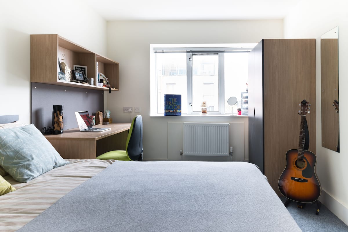 Ett sovrum med en säng, ett skrivbord, en stol, en gitarr och ett fönster med utsikt över omgivningen.