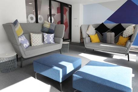 A modern office lounge with two grey sofas, patterned cushions, and blue ottomans.