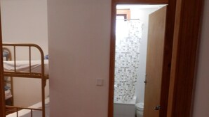 Un estrecho pasillo con una cama litera a la izquierda y un baño con inodoro y ducha al fondo.