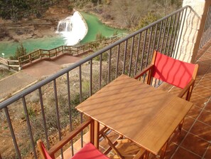 Un balcón con una mesa de madera y sillas rojas con vistas a un paisaje natural que incluye una cascada.