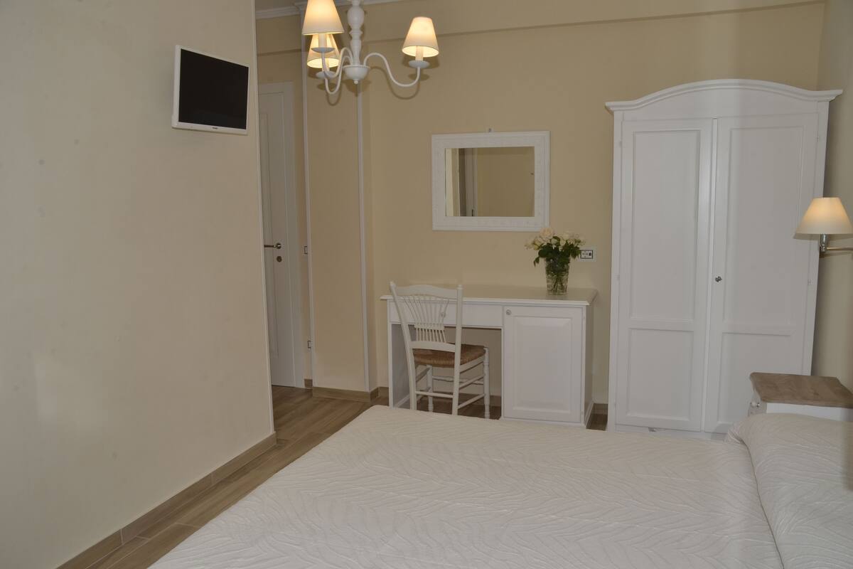 A bedroom with a bed, a desk, a chair, a wardrobe, and a chandelier.