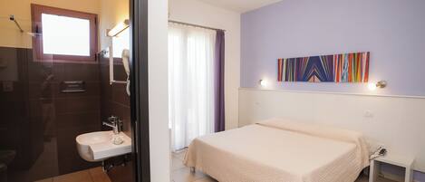 A bedroom with a bed, a bedside table, a nightstand, a window with curtains, and a bathroom with a sink and mirror.