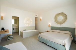 A modern bedroom with a large bed, a built-in bathtub, a vanity with a mirror, and a small stool.