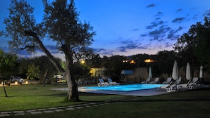 Piscina con sillones de descanso y sombrillas, rodeada de árboles y un cielo despejado.