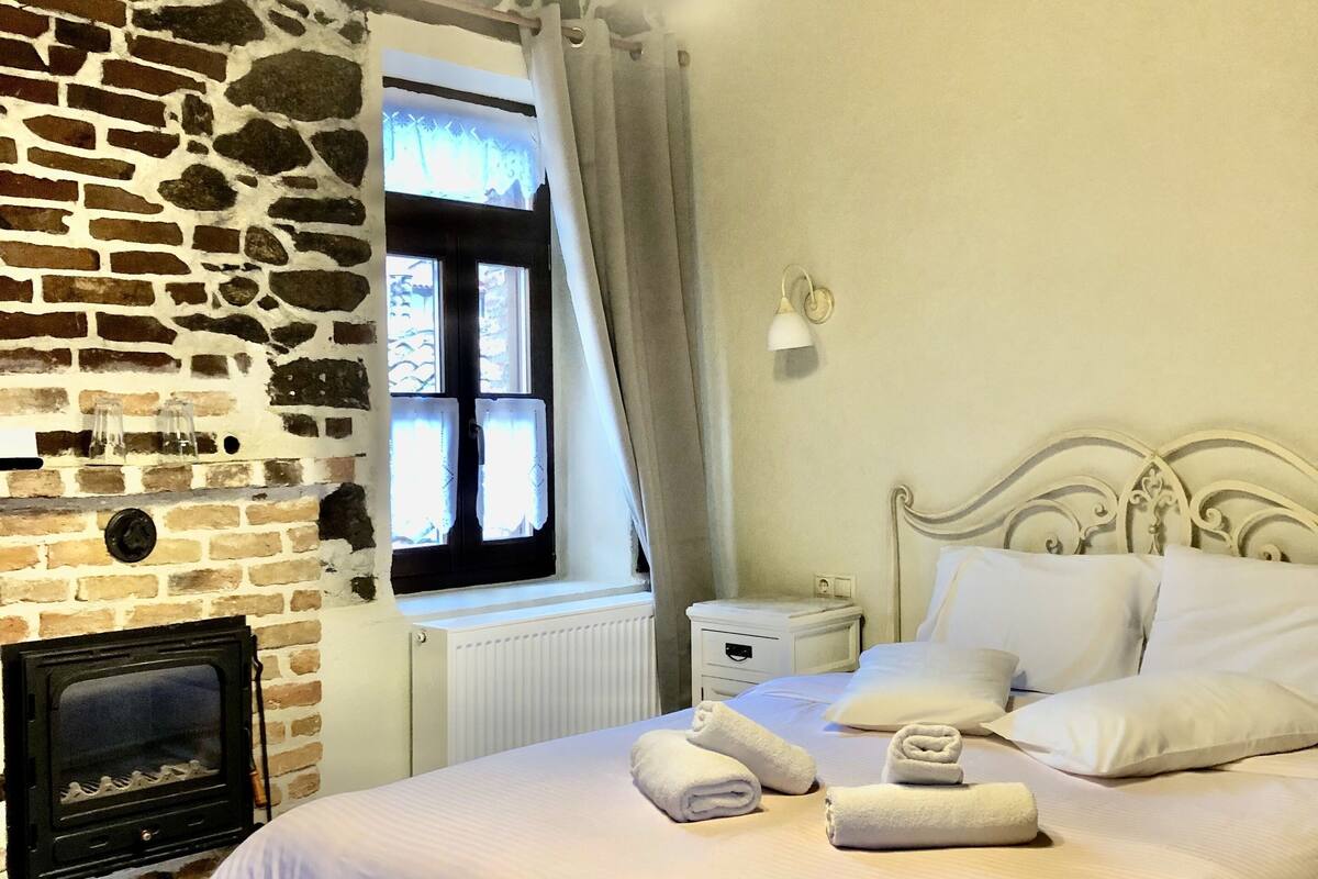 Un dormitorio con pared de piedra, chimenea, cama con ropa blanca y una mesita de noche.