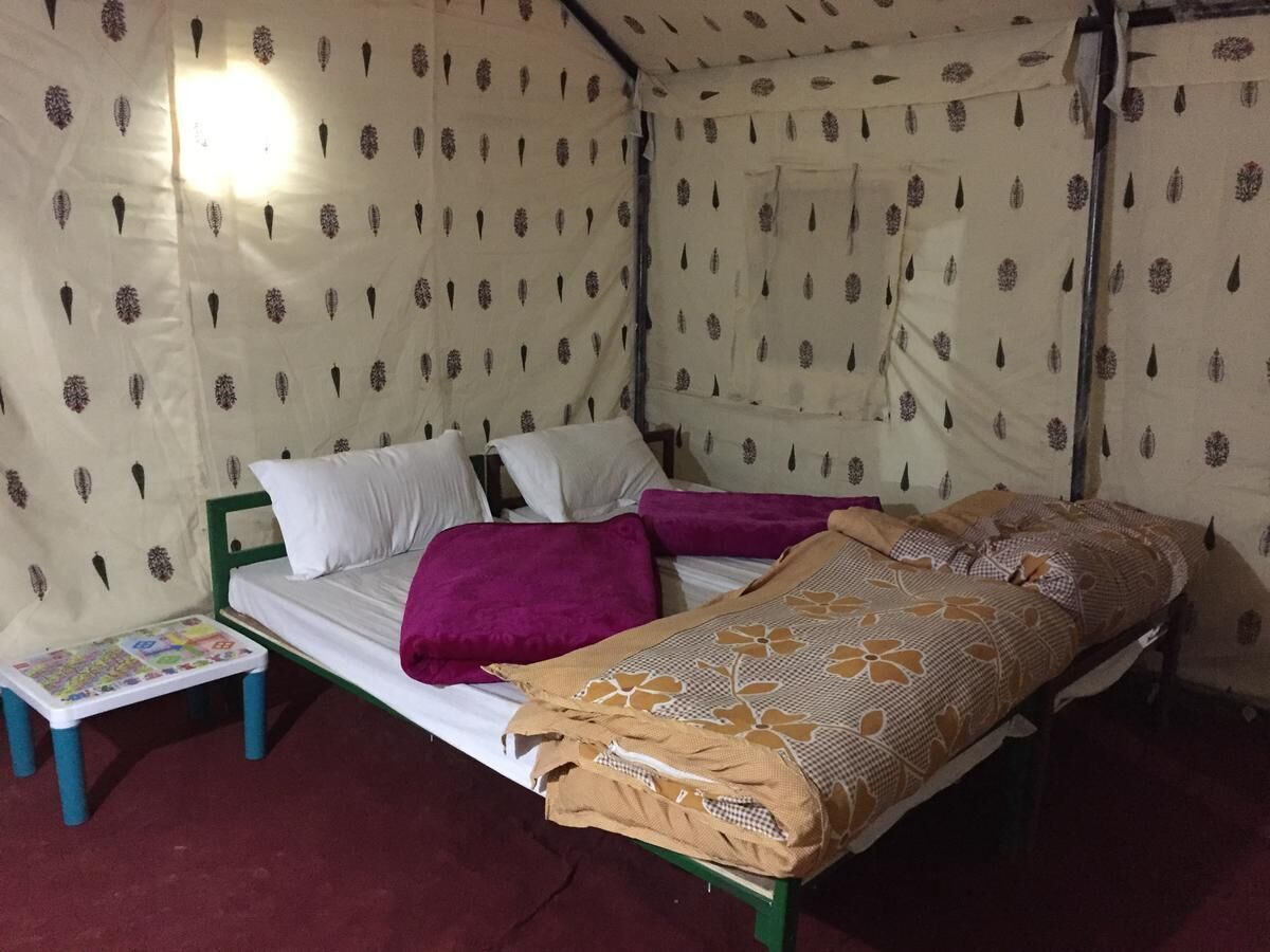 A single bed with a patterned bedspread, a purple blanket, and white pillows. There is a small table with a colorful book or magazine. The room has patterned walls and a light fixture on the ceiling.
