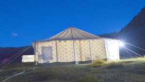A large, single-story tent with a peaked roof, situated in a grassy field with mountains in the background. - Himalayan Routes Camp (Lahul)