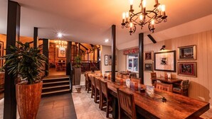 Une salle à manger spacieuse avec une longue table en bois, de nombreuses chaises et un lustre. Les murs sont ornés de tableaux encadrés, et un escalier mène à une autre pièce.
