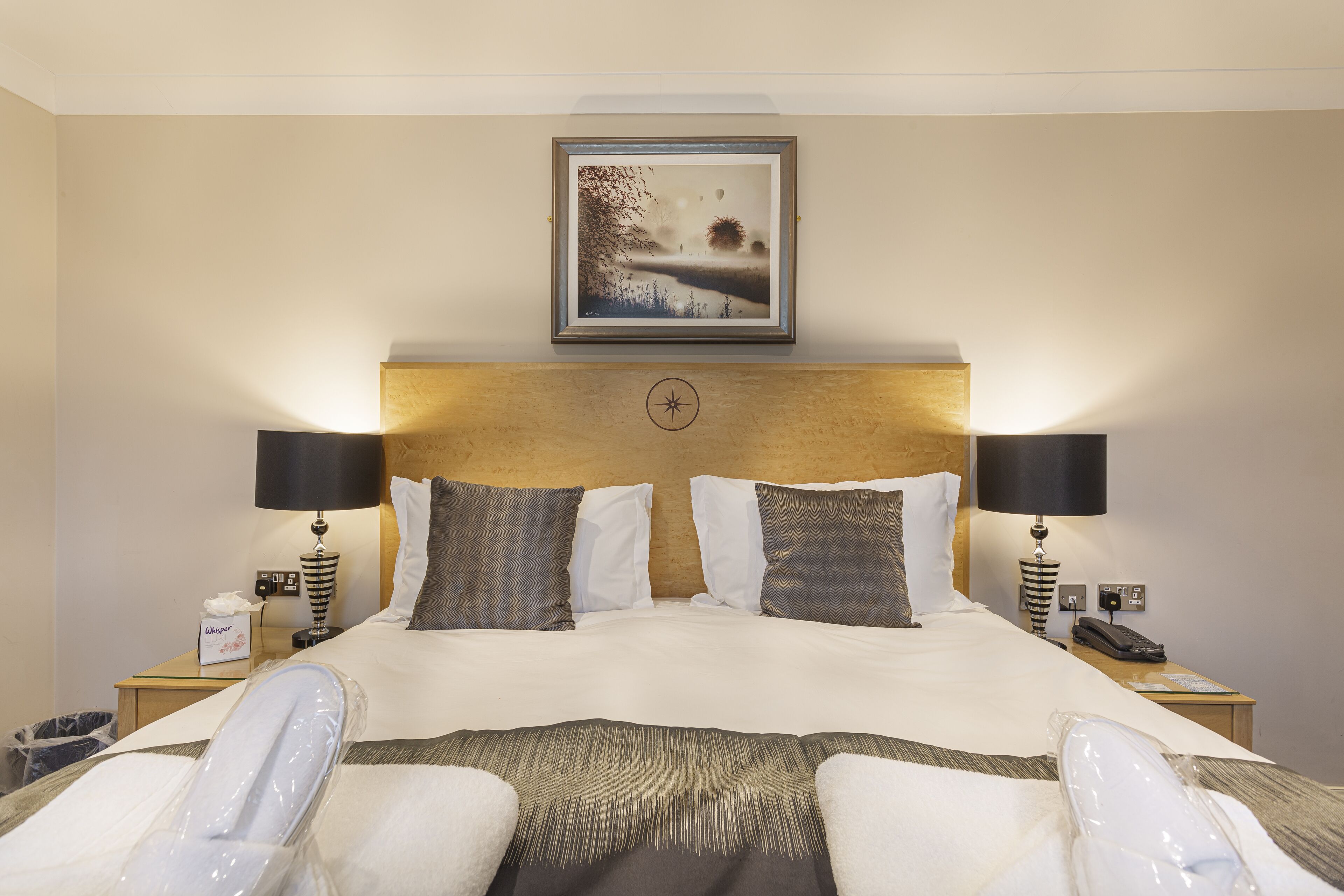 A hotel room with a large bed, two bedside lamps, a nightstand with a lamp and a phone, and a framed picture on the wall.