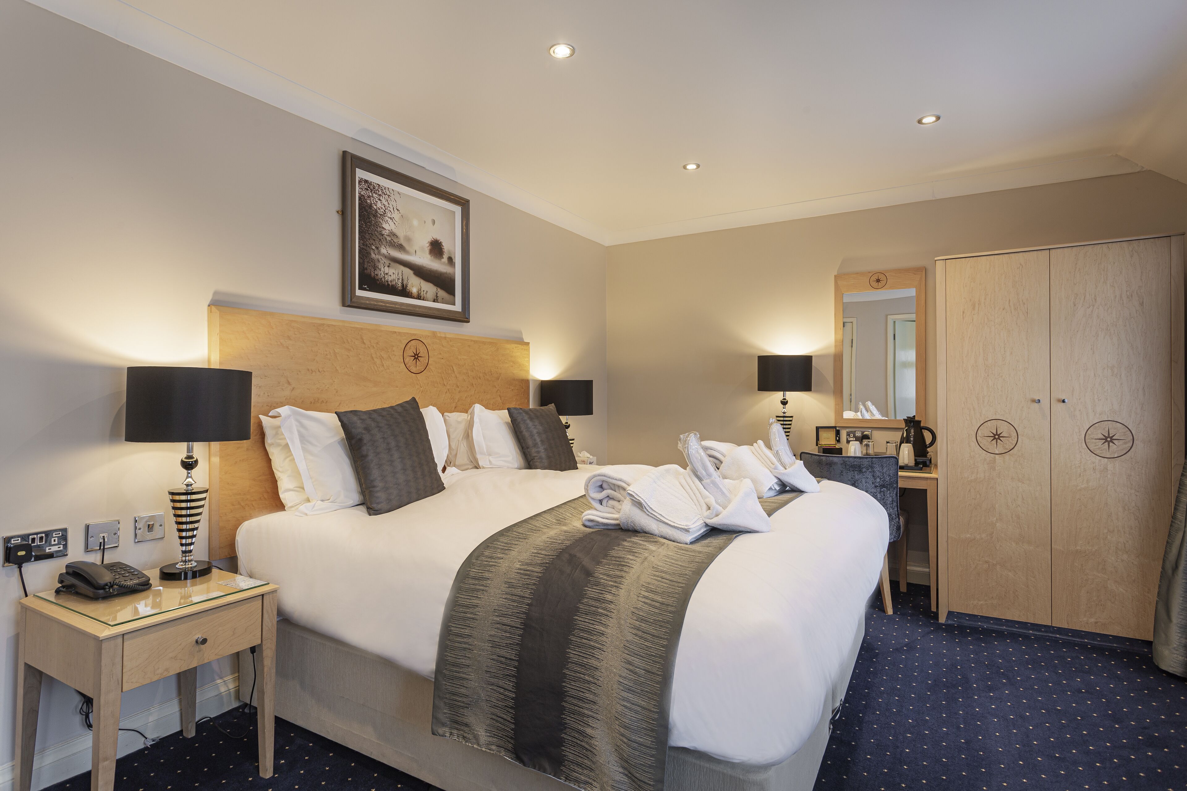 A hotel room with a large bed, a bedside table with a lamp, a desk with a mirror, and a wardrobe.