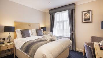 A hotel room with a large bed, a desk with a chair, a window with curtains, and a framed picture on the wall.