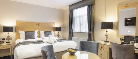 A hotel room with a bed, two chairs, a small round table with a bowl of fruit, a desk with a computer, and a large window with curtains.
