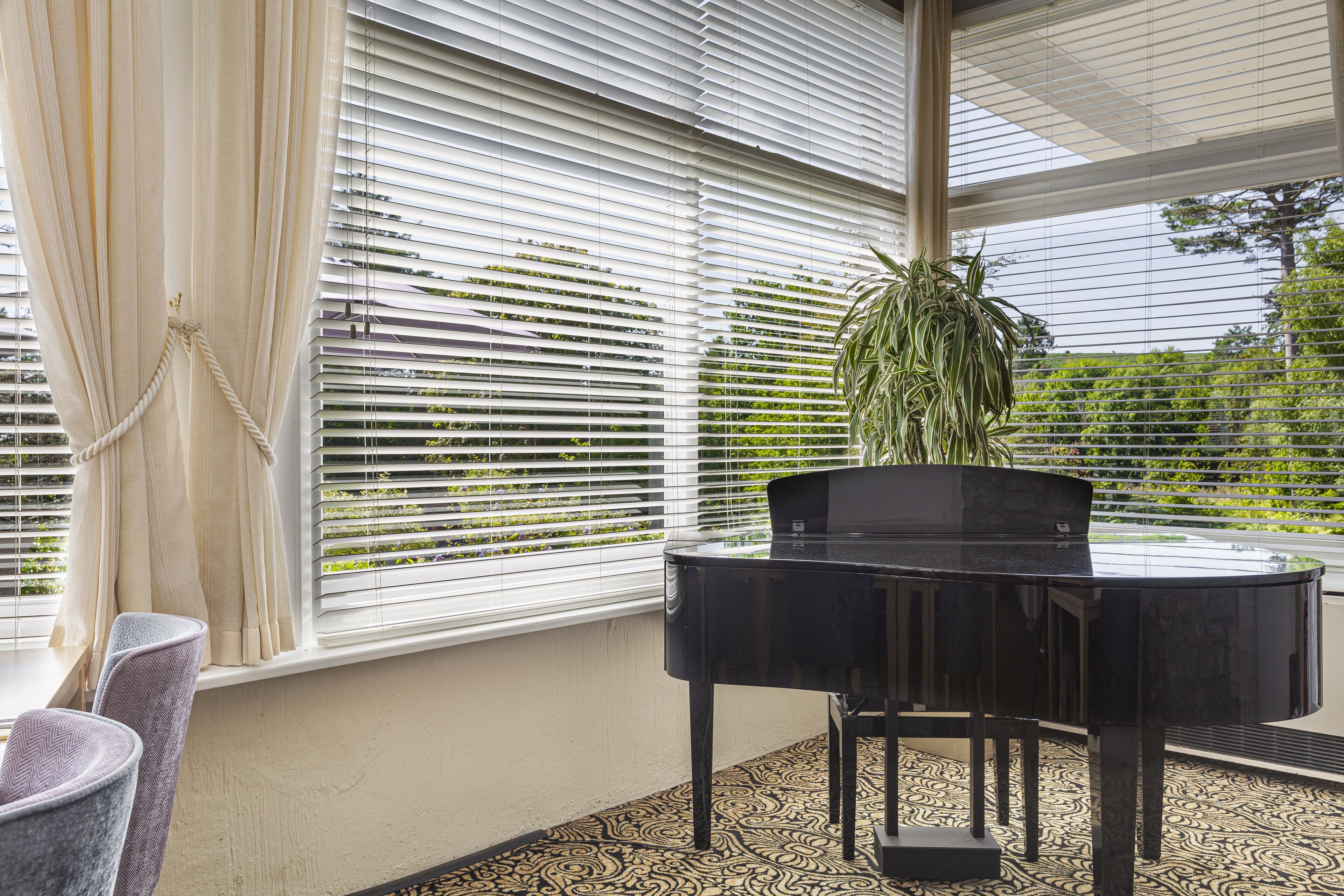 Huoneessa on piano, suuret ikkunat, ruukkukasvi ja kuviollinen lattia.