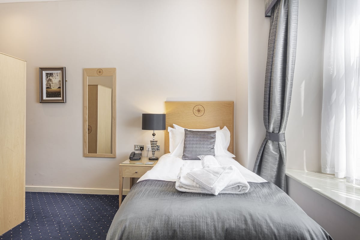 Een hotelkamer met een opgemaakt bed, een nachtkastje met een lamp, een spiegel en een raam met gordijnen.