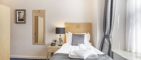 Une chambre d’hôtel avec un lit bien fait, une table de chevet avec une lampe, un miroir et une fenêtre avec des rideaux.