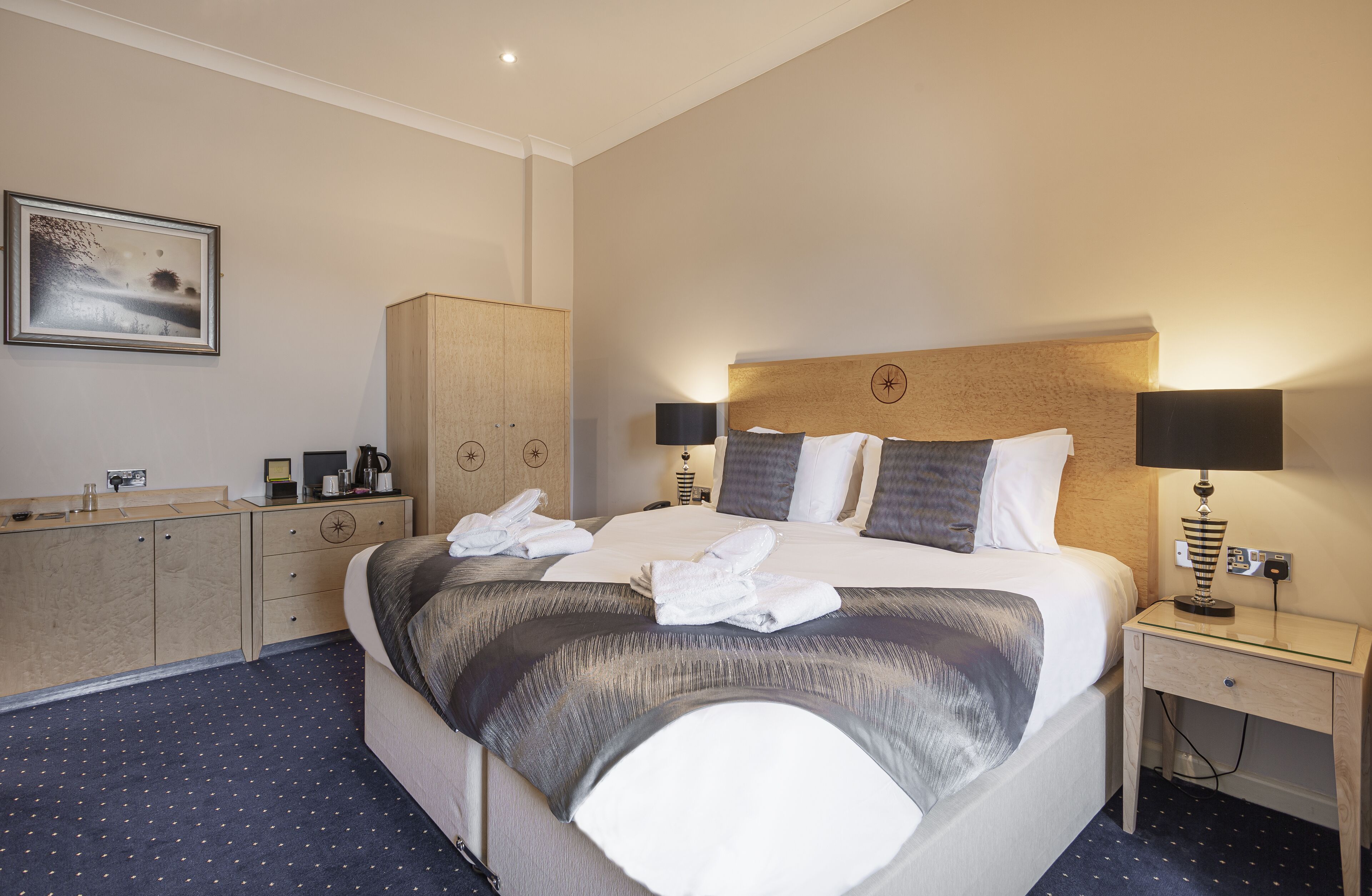A hotel room with a large bed, two bedside tables with lamps, a wardrobe, a desk with a coffee maker, and a framed picture on the wall.