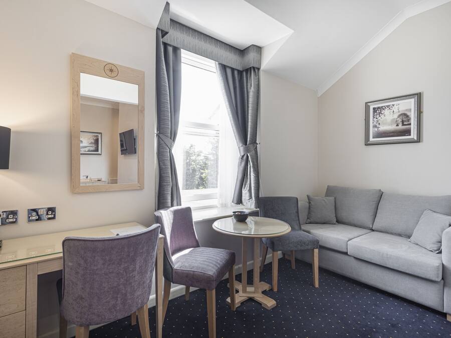Ein modernes Zimmer mit grauem Sofa, zwei Stühlen, einem kleinen runden Tisch, einem Schreibtisch mit Computer und einem Fenster mit Vorhängen.
