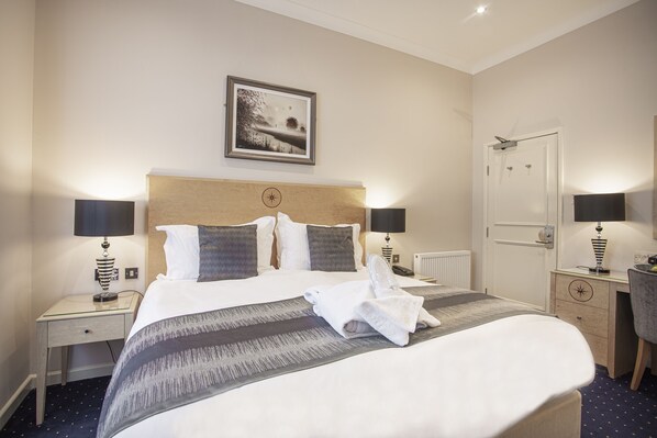 A neatly made bed with a striped blanket, two bedside lamps, a framed picture above the bed, and a small desk with a lamp to the right. - The Moorland Hotel, Haytor, Devon (Newton Abbot)
