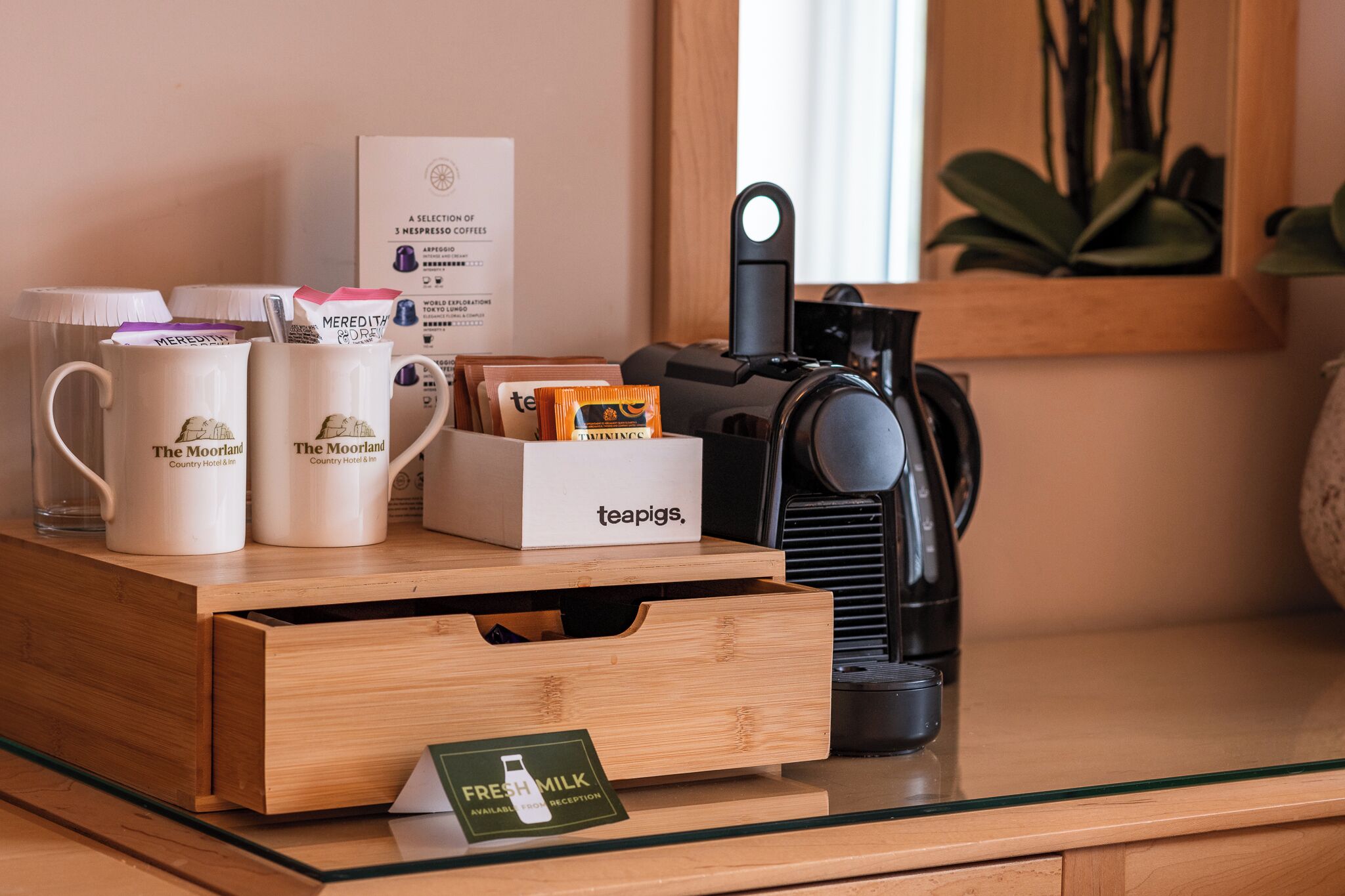 A coffee station with branded mugs, a teapig's box, a fresh milk sign, and a coffee machine on a glass table.