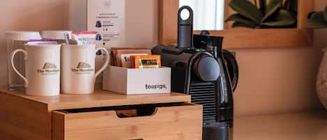 Een koffiehoek met beker met branding, een doos van Teapigs, een bord met verse melk en een koffieapparaat op een glazen tafel.
