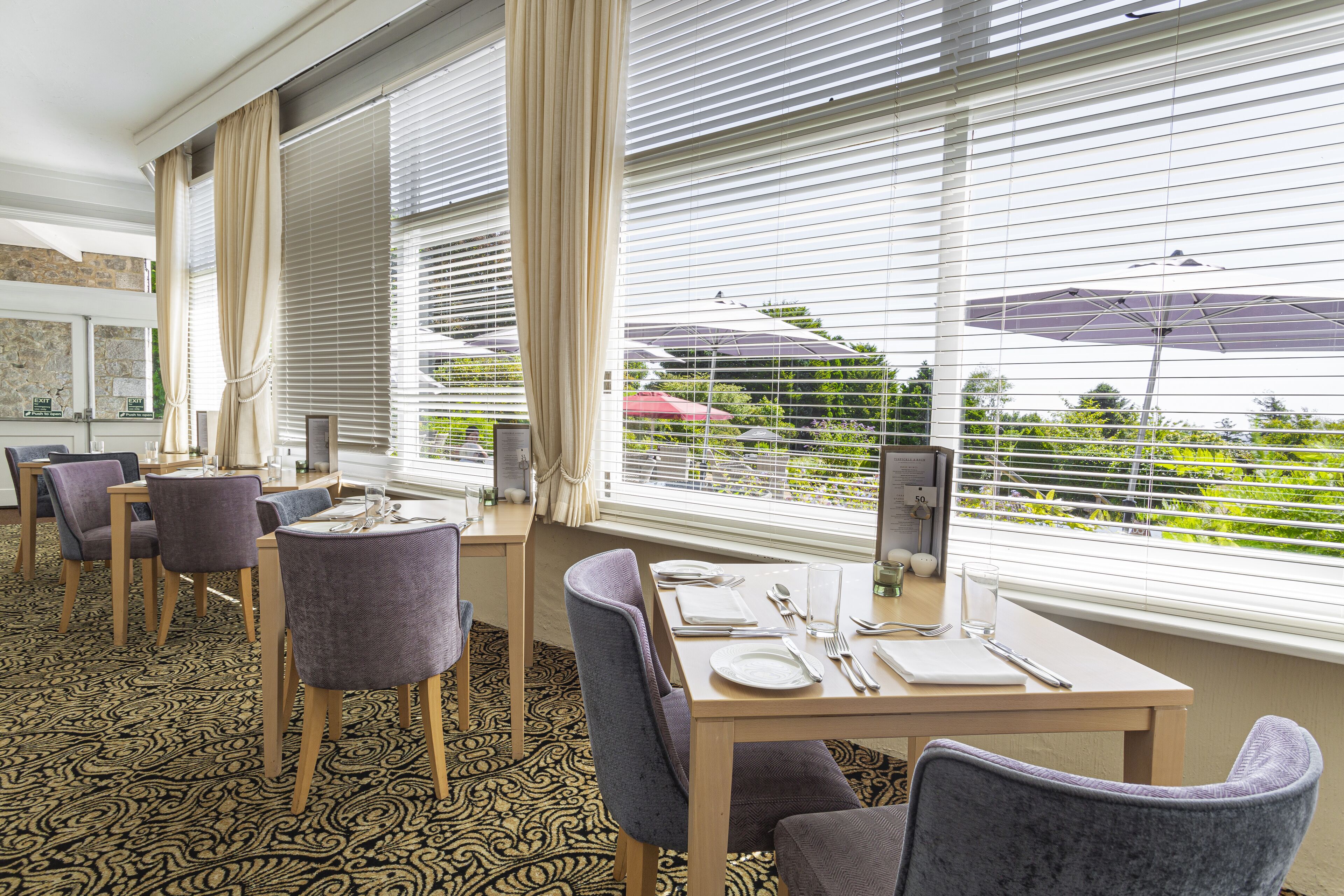 A hotel dining area with a table set for four, surrounded by chairs, large windows with blinds, and a view of greenery outside.