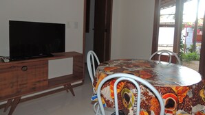 A room with a TV on a wooden cabinet, a round table with a coffee-themed tablecloth, and a door leading outside. - Nossa Casa em Jeri (Jijoca de Jericoacoara)