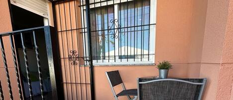 Un balcón con reja metálica, una ventana con barrotes, una silla plegable y una planta en maceta.