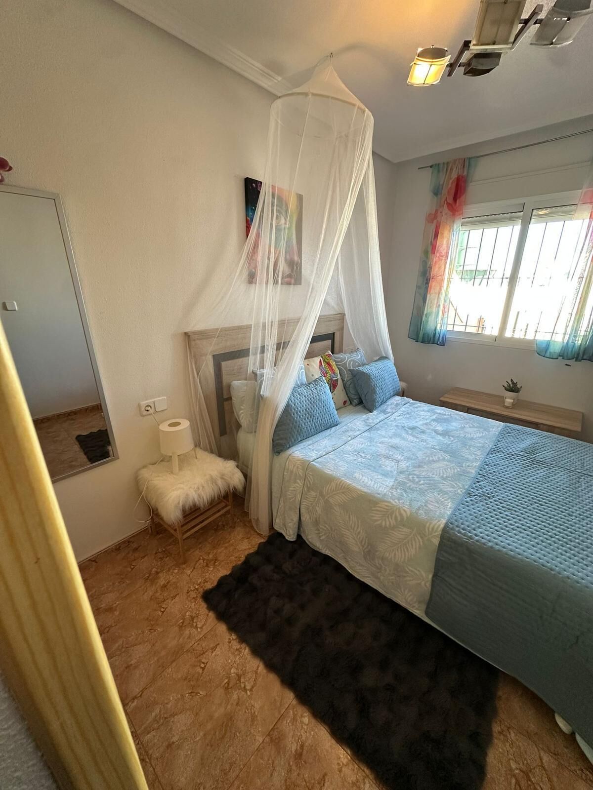 Un dormitorio con cama con dosel, un espejo, una mesita de noche con lámpara y una ventana con cortinas.