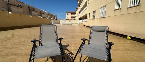 An outdoor terrace with two foldable chairs, a tiled floor, and a building with balconies in the background.