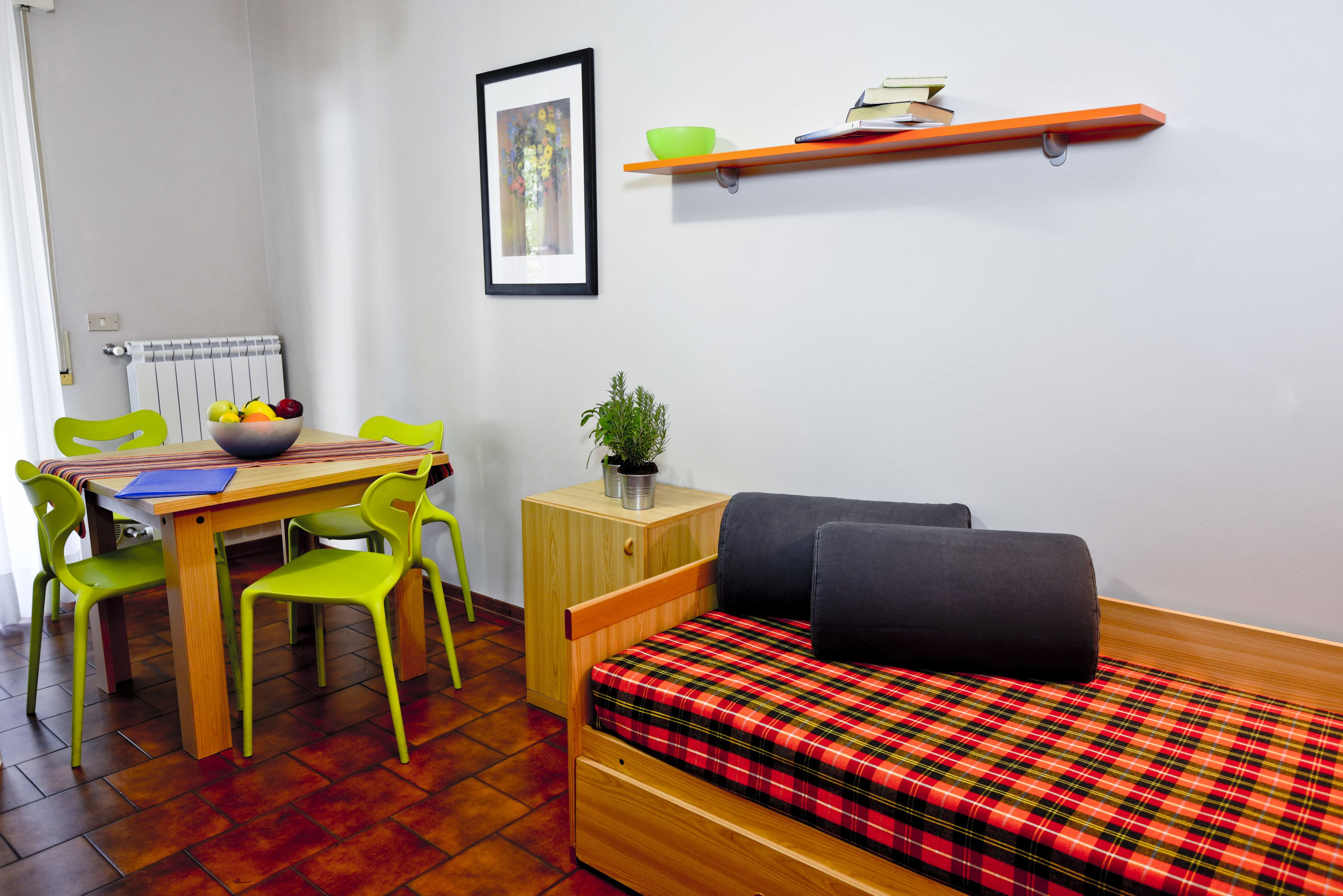 Una habitación compacta con una cama de madera, una pequeña mesa con sillas, un radiador, un cuadro enmarcado, un estante con libros y un cuenco, y una planta en maceta.