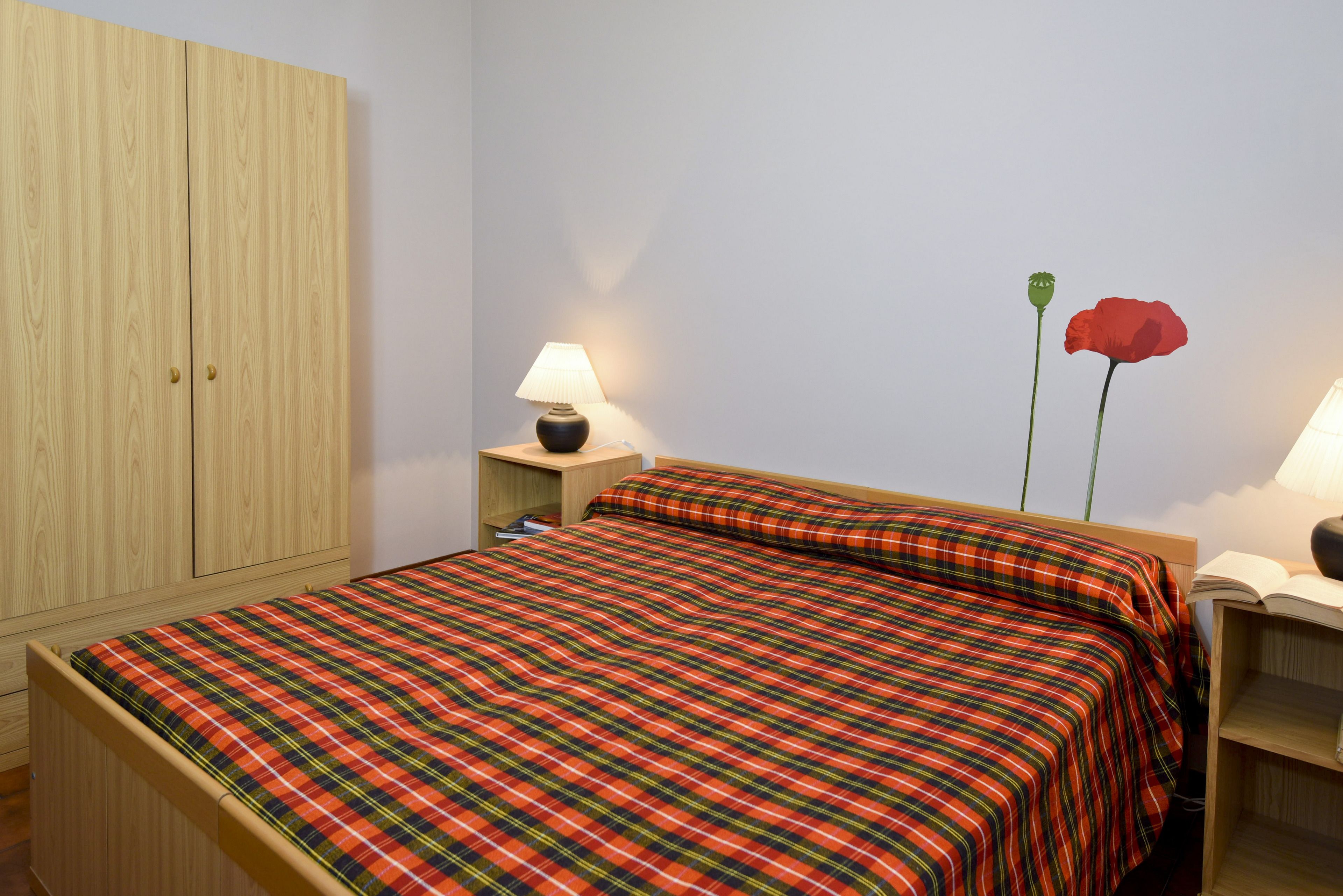 A bedroom with a plaid bedspread, a wooden wardrobe, a bedside table with a lamp, and a single red flower on the nightstand.