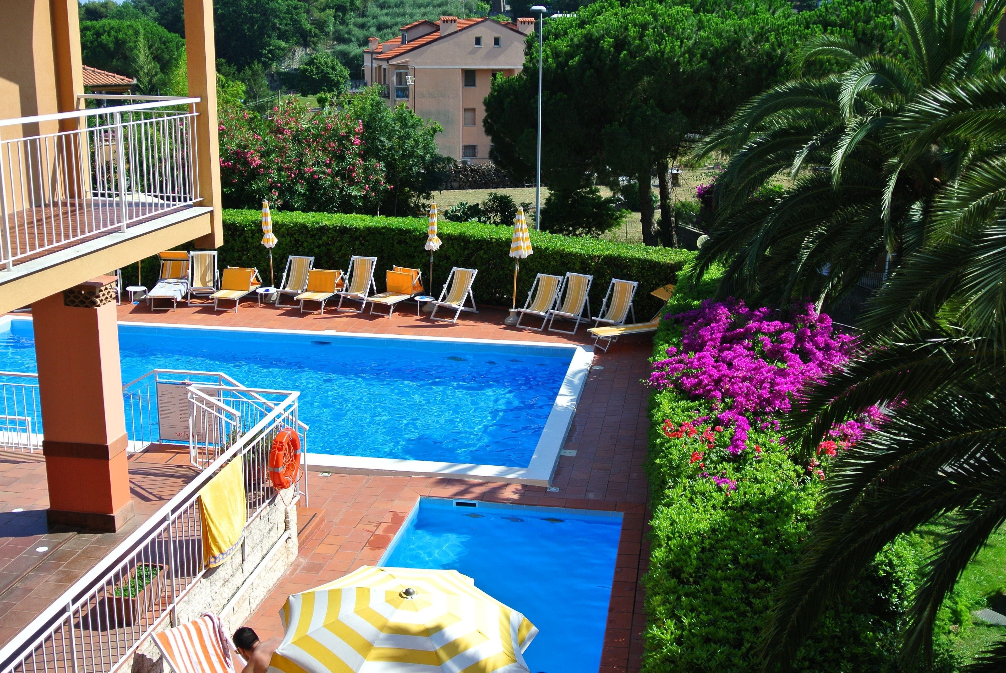 A swimming pool with sun loungers and umbrellas, surrounded by lush greenery and a building in the background.