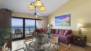 Ein Wohnzimmer mit Glastisch, Stühlen, einem violetten Sofa, einem Sonnenuntergangsgemälde und Blick auf den Strand durch Balkontüren.