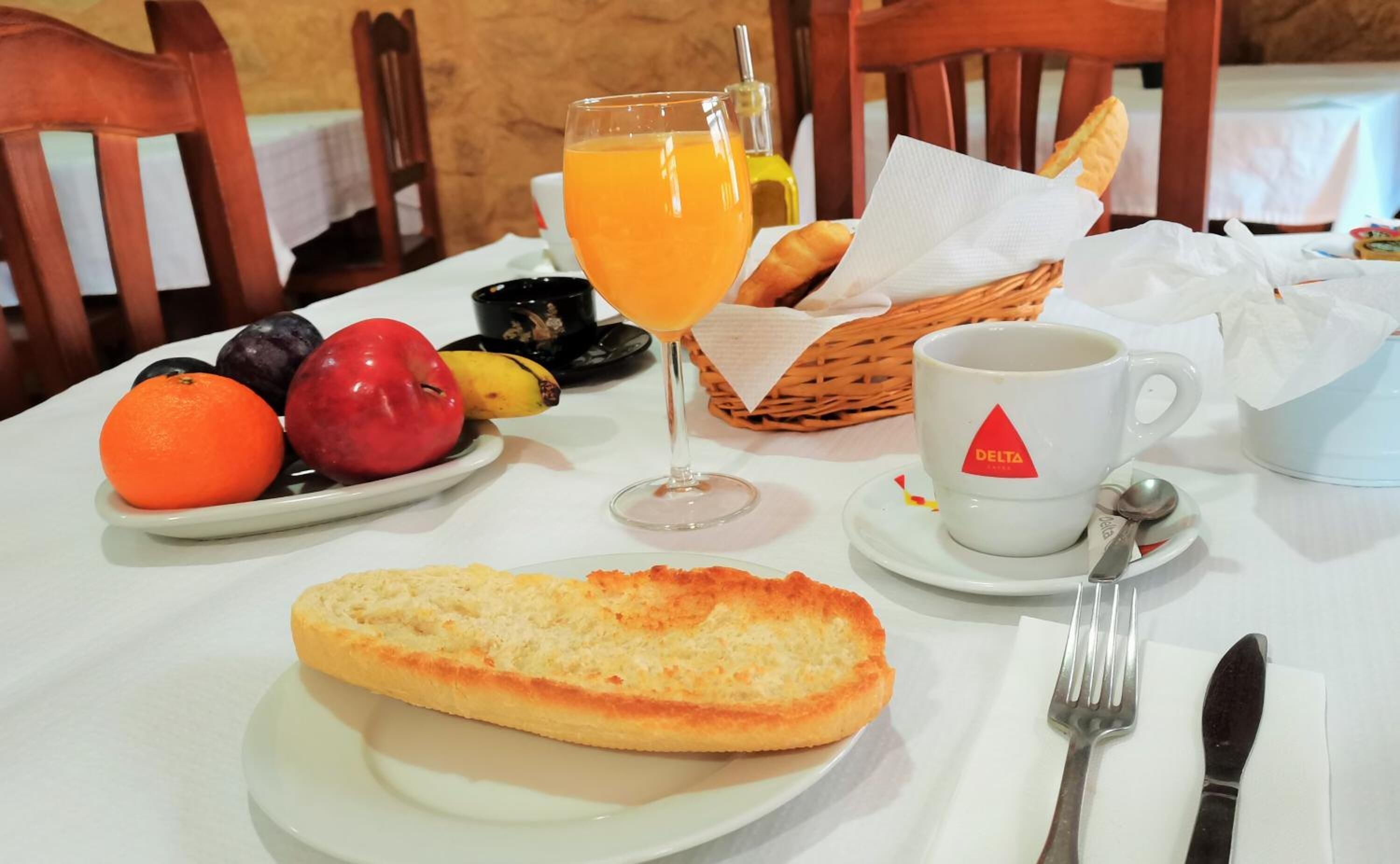 A breakfast setting with a plate of fruit, a glass of orange juice, a slice of toast, a cup of coffee, and a basket of bread.
