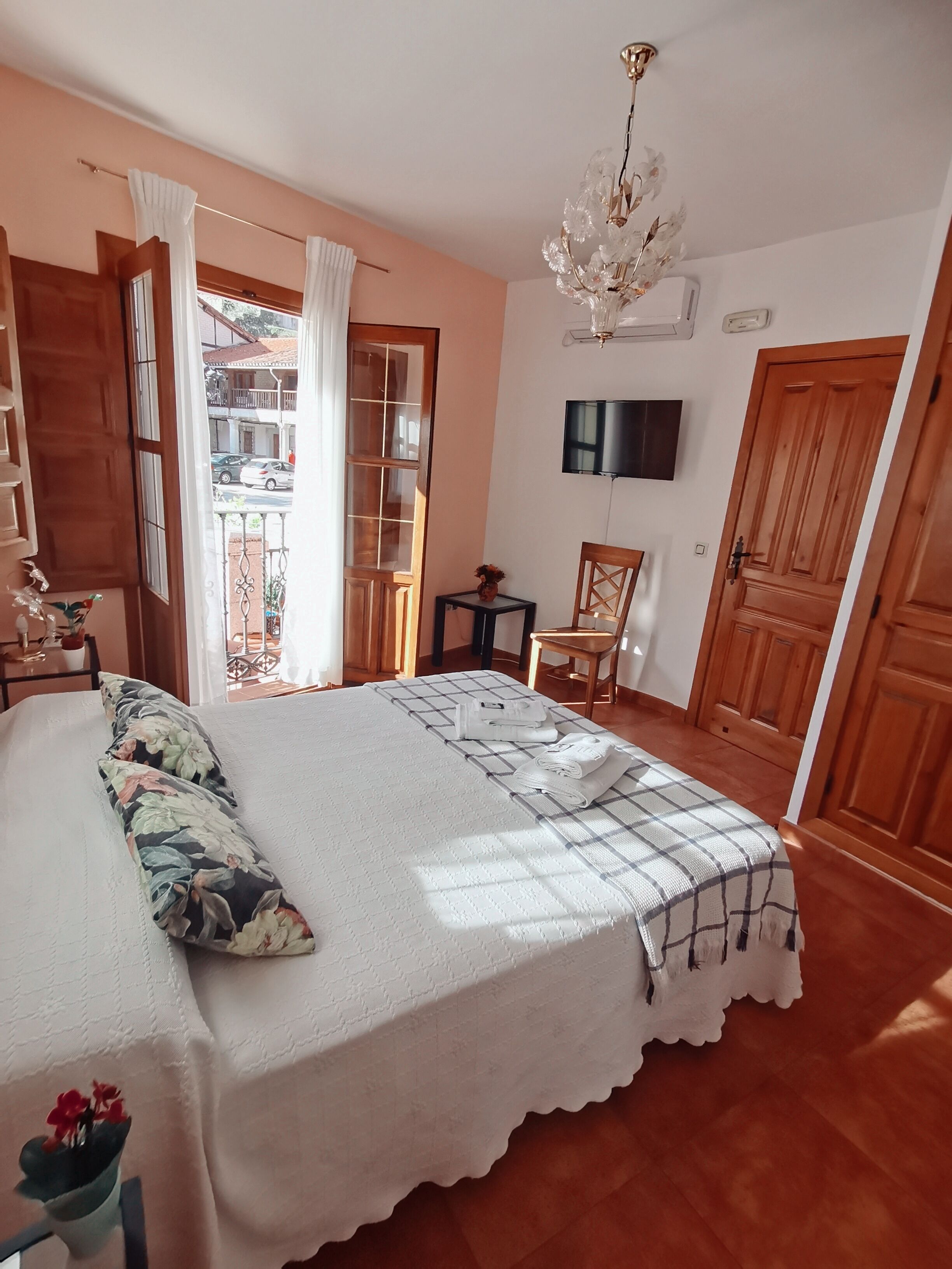 A bedroom with a large bed, wooden flooring, a chandelier, a TV, and a view of the street through a window.