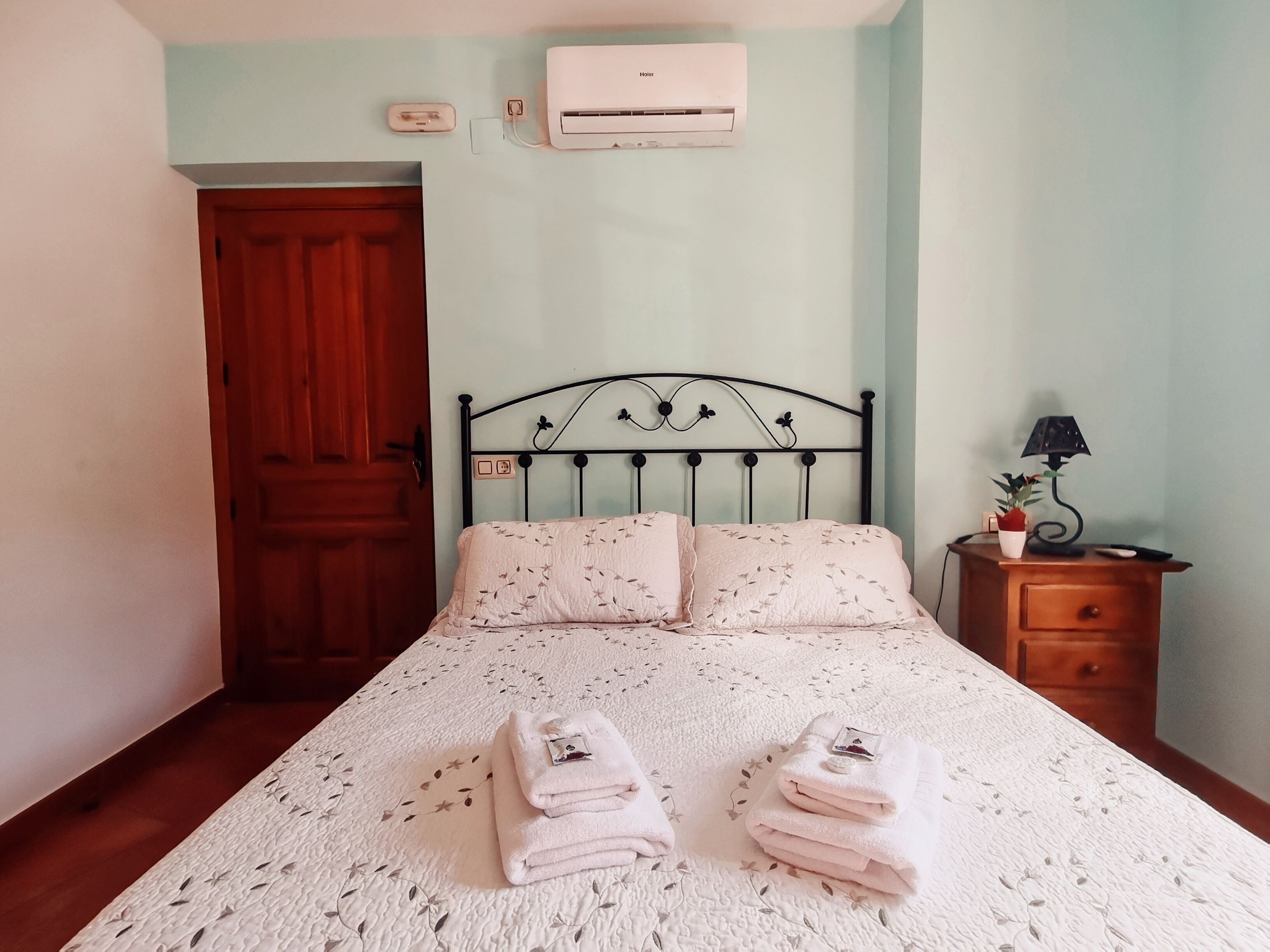 Een net opgemaakt bed met een bloemenprintdeken en kussens, een houten nachtkastje met een lamp en een potplant, en een airconditioner aan de muur.