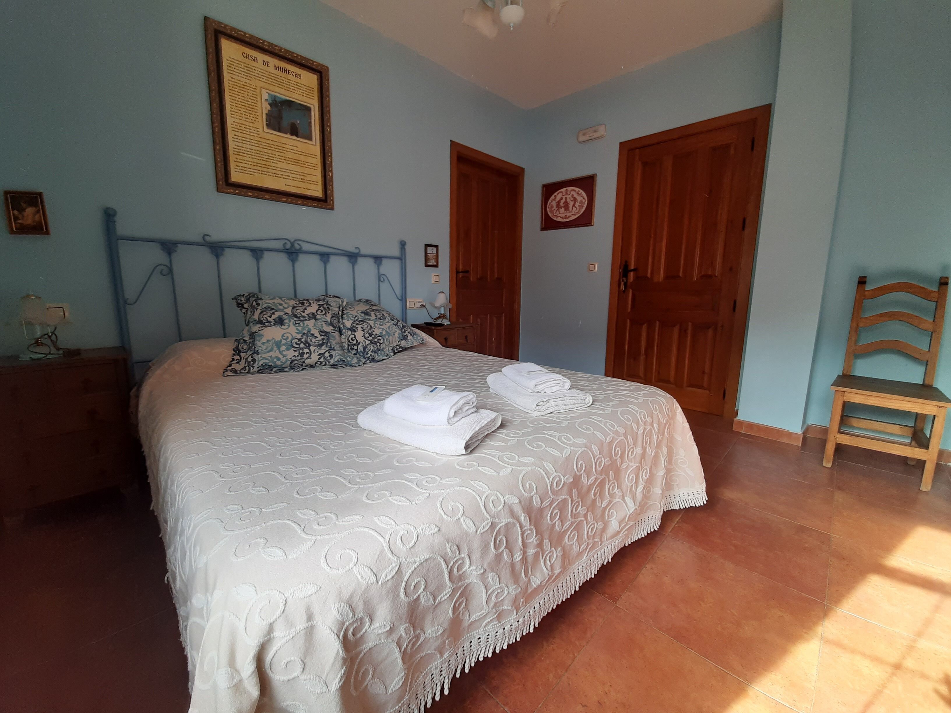 A bedroom with a bed, a wooden chair, a nightstand, a framed picture on the wall, and a door.