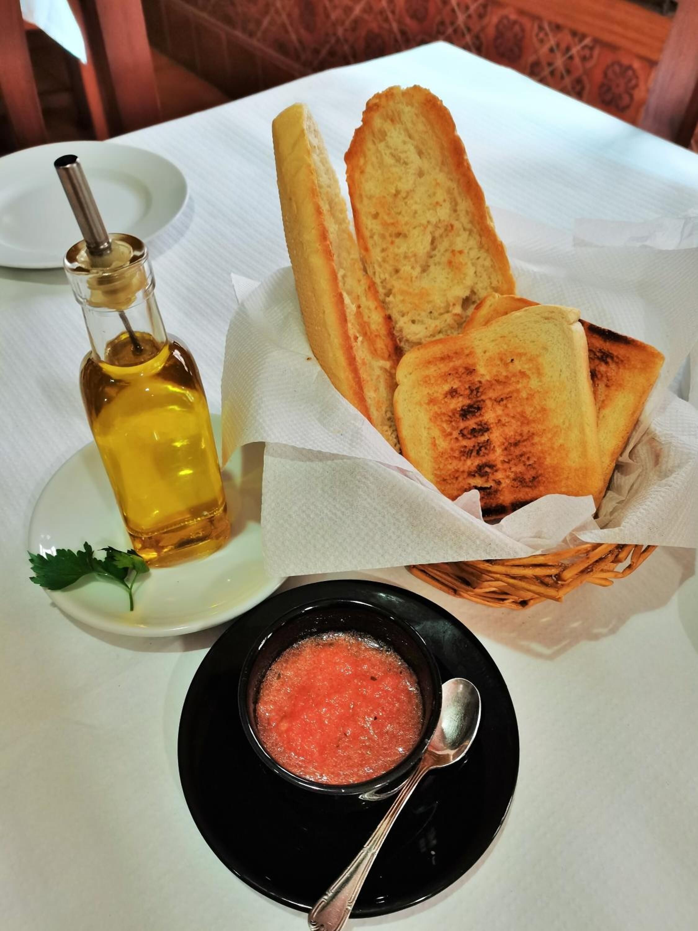 Une table avec un panier de pain grillé, une bouteille d’huile d’olive et un bol de soupe à la tomate.