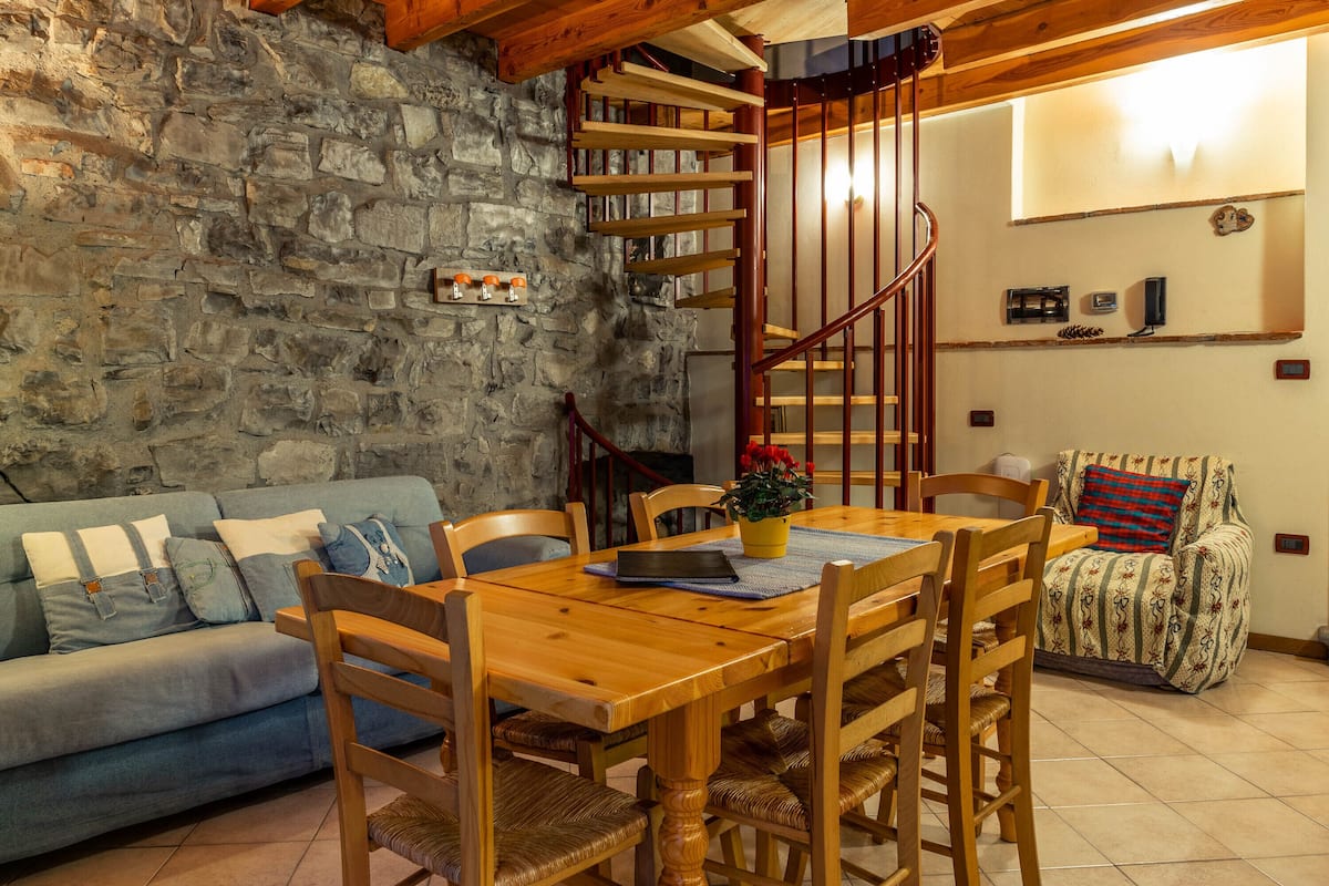 Una sala de estar acogedora con una pared de piedra, una escalera de madera, un sofá azul, una mesa de comedor de madera con sillas y un sillón con estampado.