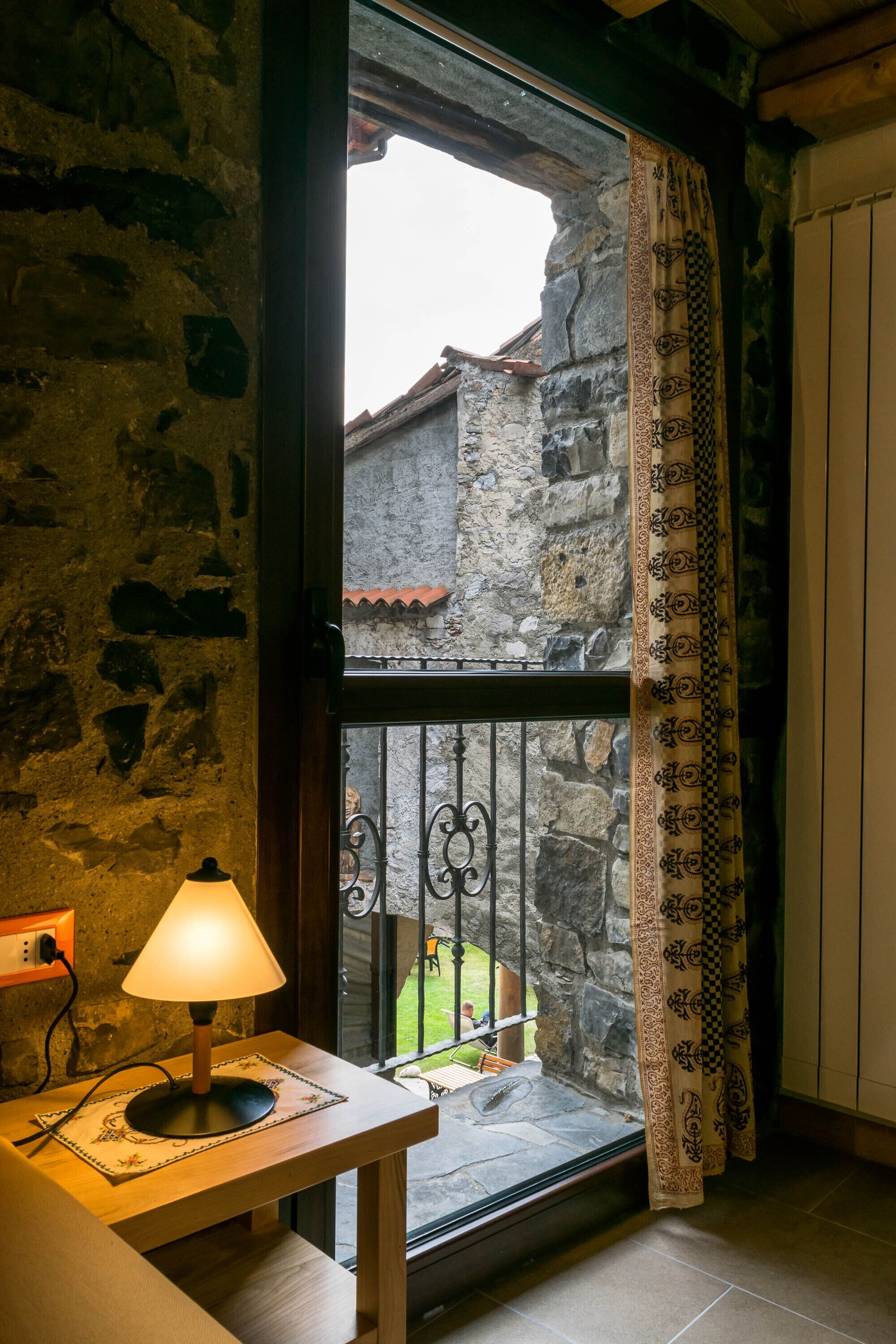 A stone wall, a wooden table with a lamp, a window with a metal gate, and a patterned curtain.