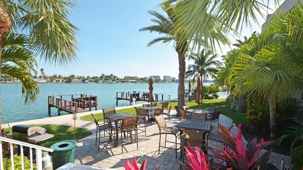An outdoor patio with tables and chairs overlooking a body of water, surrounded by palm trees and greenery. - Come make a snowman out of sand in warm, sunny Florida! (Treasure Island)