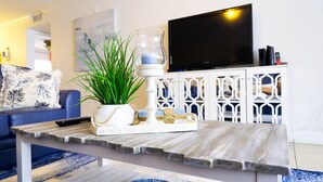 A living room with a wooden coffee table, a potted plant, a white decorative tray, and a flat-screen TV on a white cabinet. - Come make a snowman out of sand in warm, sunny Florida! (Treasure Island)